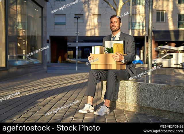 Smiling businessman with box sitting on bench at footpath
