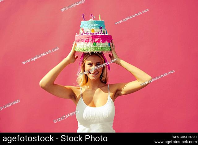 Smiling woman carrying birthday cake on head against pink wall
