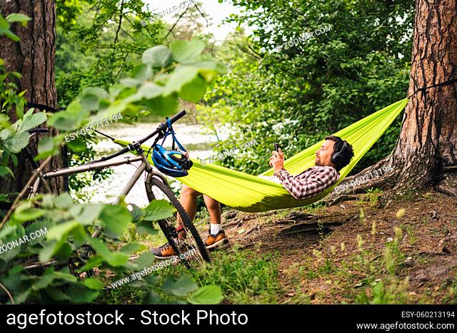 Young caucasian man resting in hammock, listening to music on headphones and using smartphone afterwards on bicycle in forest near the lake