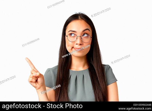 Skeptical and doubtful asian female in glasses, smirk as looking and pointing finger at upper left corner with hesitating expression, having doubts