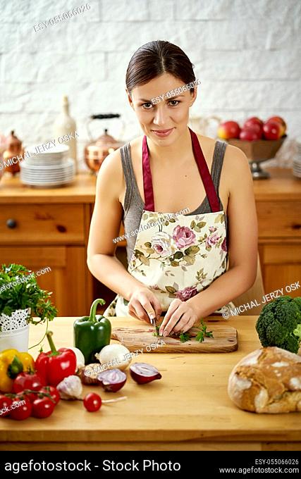beautiful young woman, brunette slicing parsley in the kitchen at a table full of organic vegetables