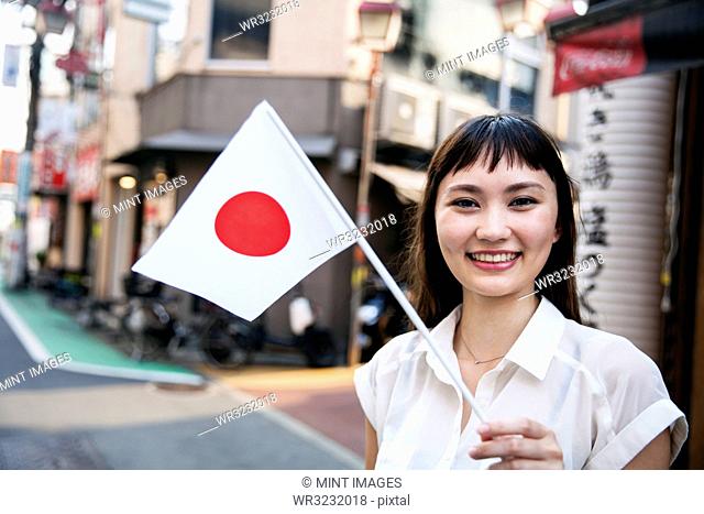 Smiling Japanese woman with long brown hair wearing white short-sleeved blouse standing in a street, holding small Japanese flag