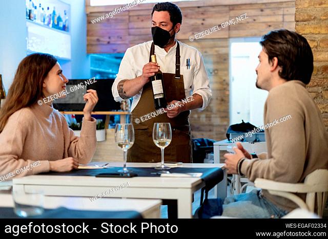 Couple talking with waiter holding wine bottle at restaurant during COVID-19