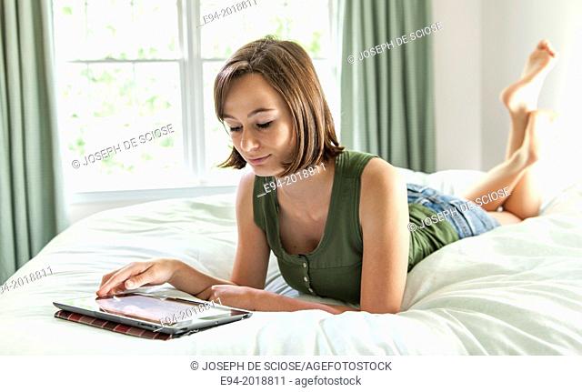 An 18 year old girl lying on her bed reading from a pad computer,