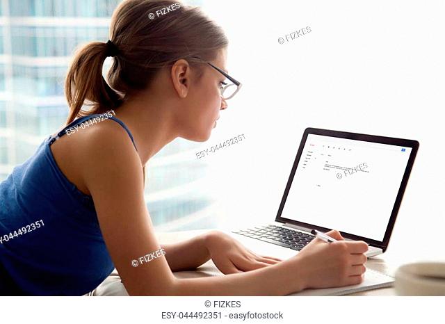 Young serious woman in glasses looks at laptop screen, writing notes. Girl composing email reply to potential employer, university admissions