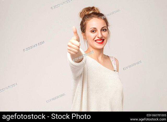 Young adult woman thumbs up, looking at camera with toothy smile. Studio shot, isolated on gray background
