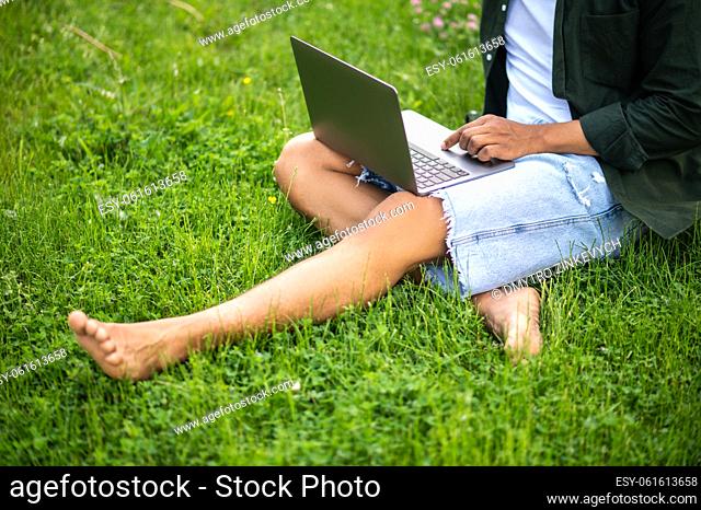 Time for yourself. Legs of dark-skinned man working on laptop sitting on grass in nature without face