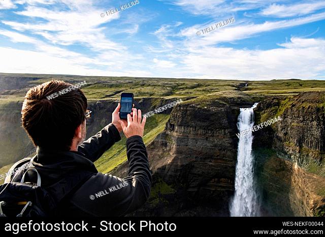 Tourist taking photo of Haifoss waterfall through mobile phone while standing at Iceland