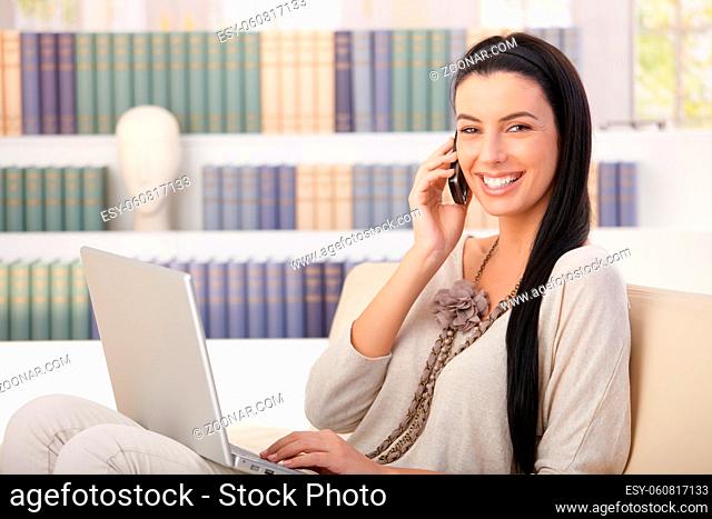 Laughing pretty woman sitting on couch at home with laptop computer, speaking on mobile phone