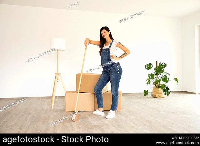 Pregnant woman with broom standing by boxes in new unfurnished house