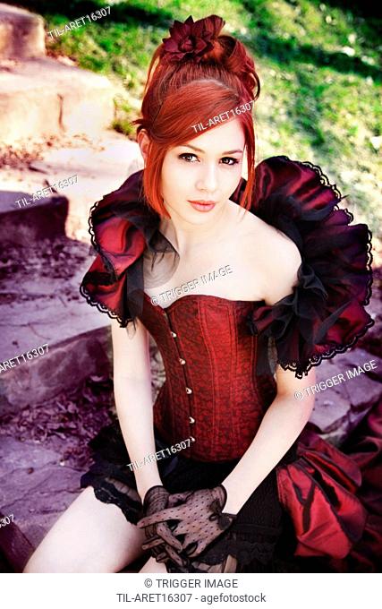 Portrait of young woman with red hair dressed in victorian style corset, sitting on stairs outdoors looking at camera