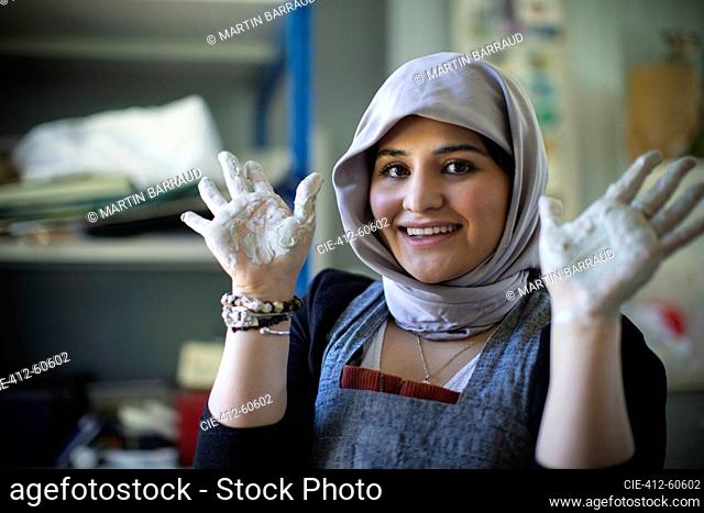 Portrait happy young female artist in hijab with clay covered hands