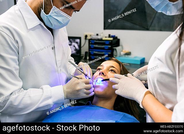 Male dentist in mask and gloves examining teeth of female patient with help of assistant at clinic