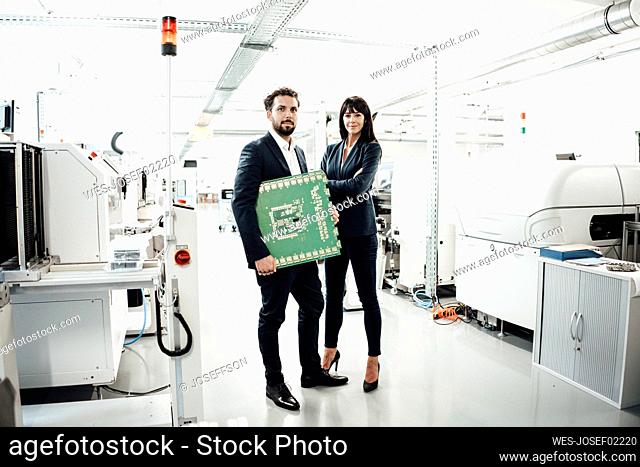 Confident businessman holding computer chip while standing by female colleague with arms crossed at industry