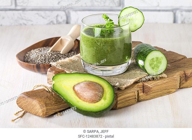 Healthy green juice smoothie surrounded by avocado, cucumber, celery and chia seeds on white background