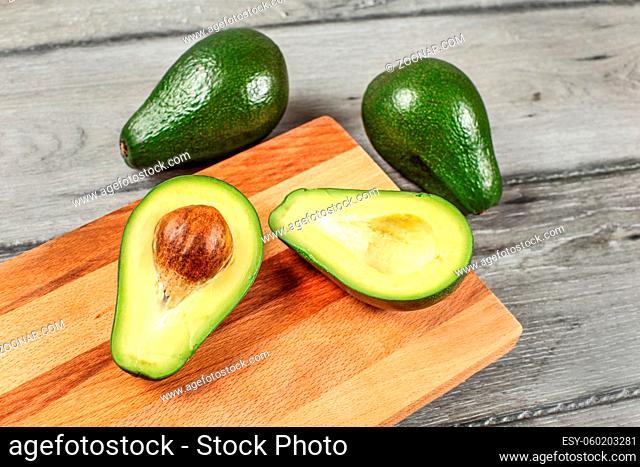 Avocado on wooden chopping board cut in half, seed visible, whole green avocado pears in background