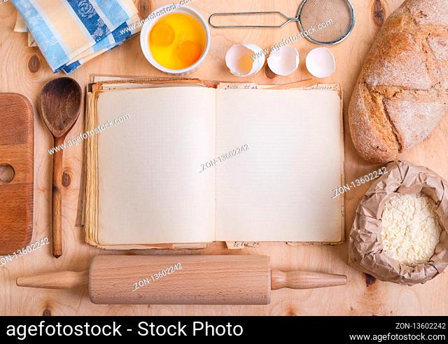Baking light warm background with blank cook book, cutting board, eggshell, bread, flour, rolling pin. Vintage wood table from above