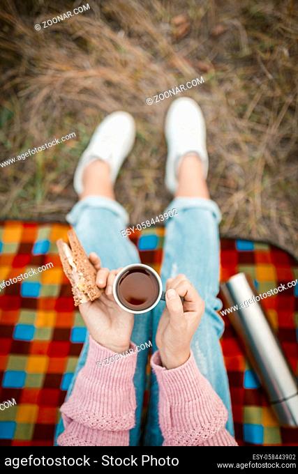 Picnic or snack in nature. Female hands holding a cup of tea and a sandwich. Young woman or teen girl snacking while sitting on grass. Close-up shot