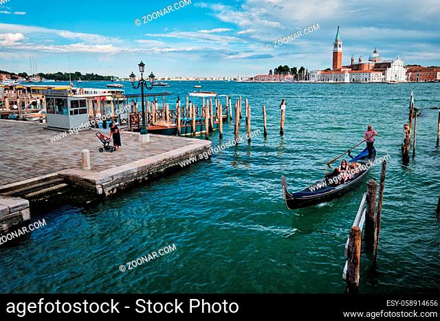 VENICE, ITALY - JUNE 27, 2018: Gondolier with tourists in gondola in lagoon of Venice by Saint Mark (San Marco) square with San Giorgio di Maggiore church in...