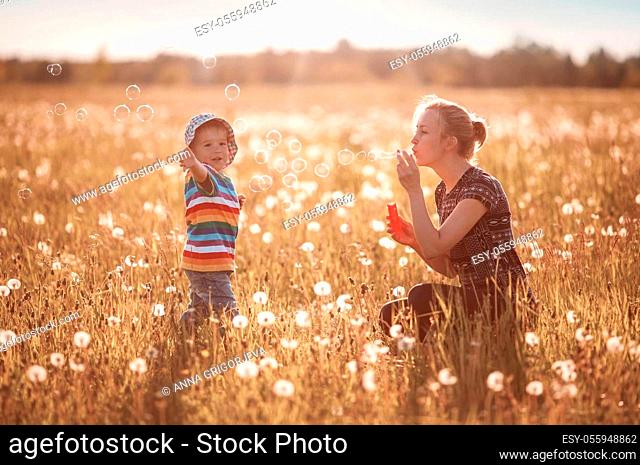 Young woman with a boy blowing bubbles. Mother and son having fun on dandelion field in nature in summer evening