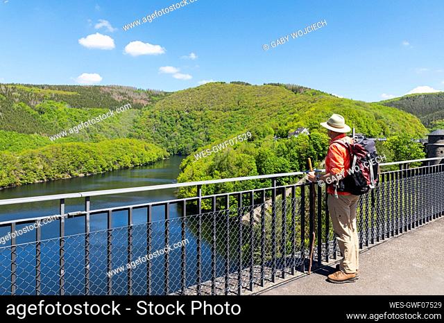 Senior man wearing hat and backpack standing by railing looking at view, Eifel National Park, Germany