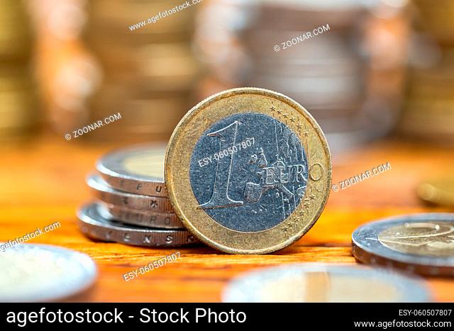 One euro coin standing on wooden table from close up. Metalic money from deatil with pile of others in background. Concept of prosperity and saving money