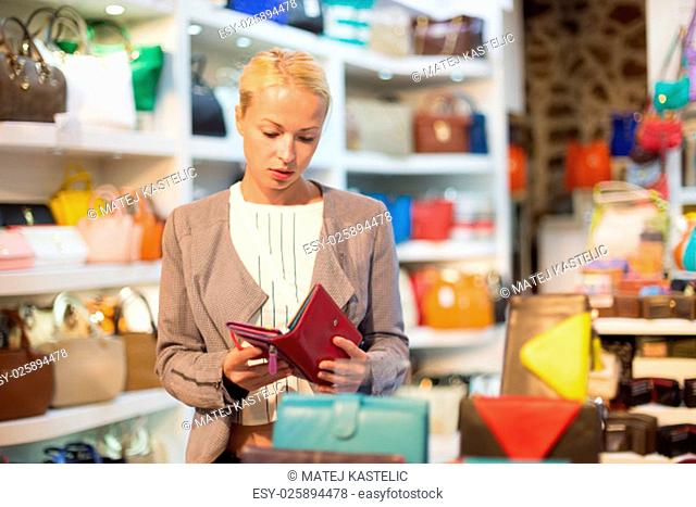 Beautiful casually dressed caucasian blond woman shopping for new colorful stylish leather wallet in fashion boutique