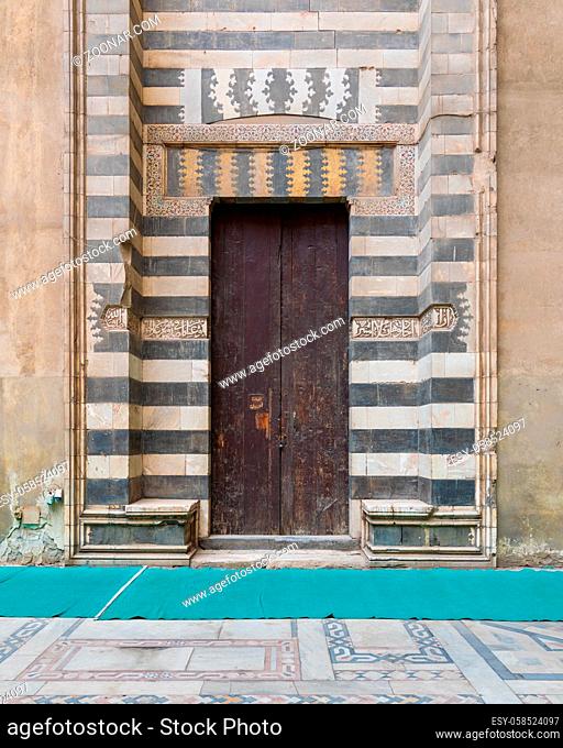 Wooden aged door surrounded by striped black and white marble decorations and stone wall at the courtyard of Sultan Hassan Mosque, Cairo, Egypt