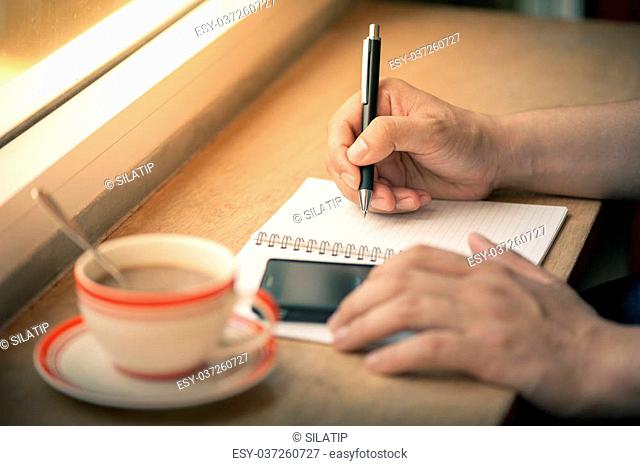 Male right hand writing on notebook with another hand using smartphone on wood bar beside window in cafe in morning time