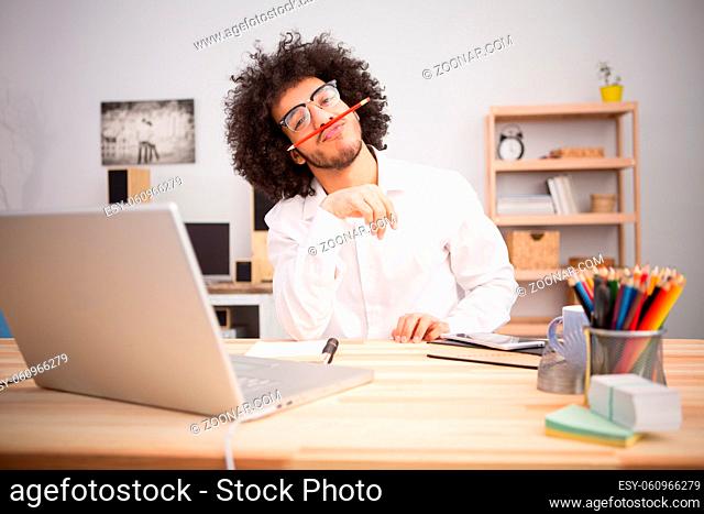 Hipster freelance man joking while working on laptop computer at home alone. Handsome man in glasses looking at camera and holding pencil between nose and mouth