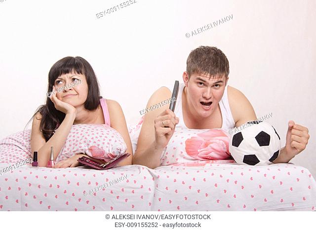 Young girl and a guy in bed. Beautiful girl has a manicure, man watches football on TV