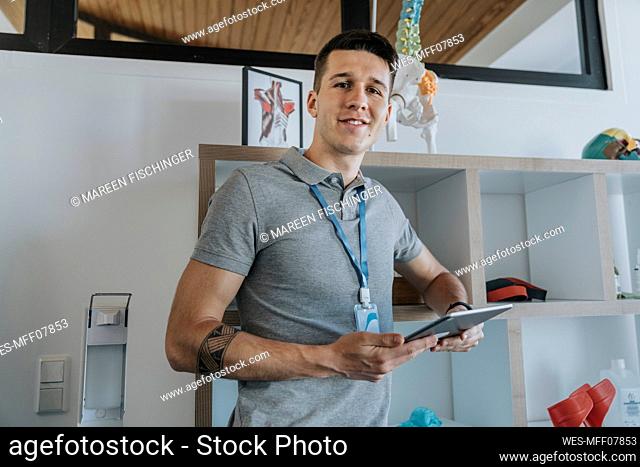 Young male physiotherapist with digital tablet standing by rack in medical practice