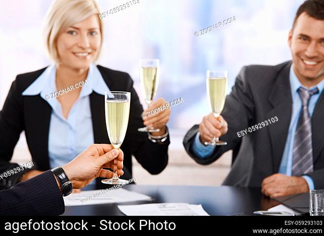 Happy young business people sitting around meeting table at office raising toast with champagne, smiling. Focus placed on hand in front