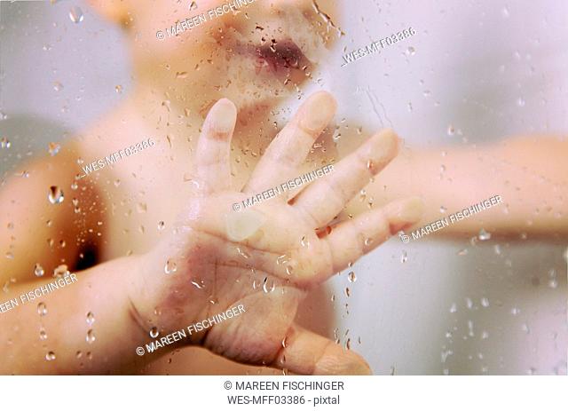 Hand of little boy against wet glass pane in shower