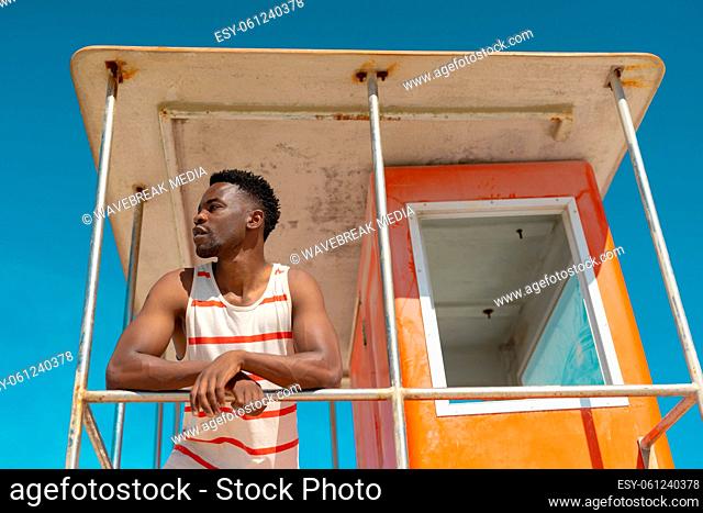 Low angle view of african american young man looking away standing on lifeguard hut against blue sky