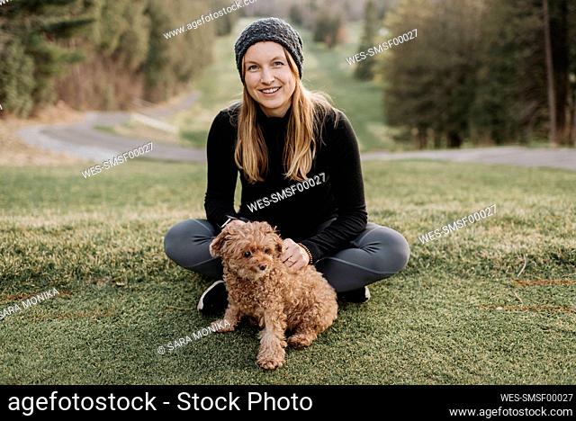 Smiling woman with poodle sitting on grassy land in park