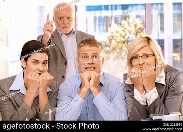 Scared employees sitting with hands up to mouth, angry boss standing behind pointing