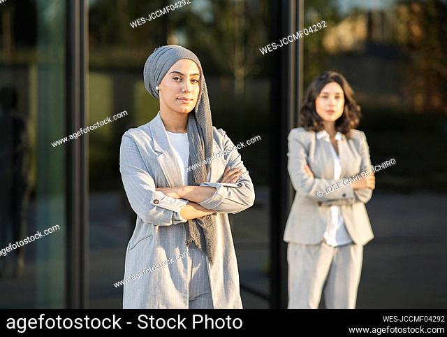 Confident businesswoman with arms crossed standing by female colleague in front of glass wall