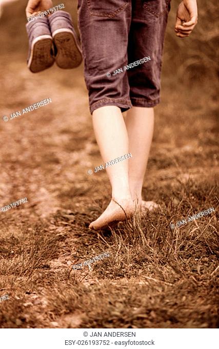 Rear close up view of unidentifiable person walking on grassy dirt path and holding shoes