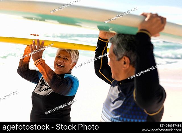 Cheerful senior biracial woman carrying surfboard over head looking at man doing the same