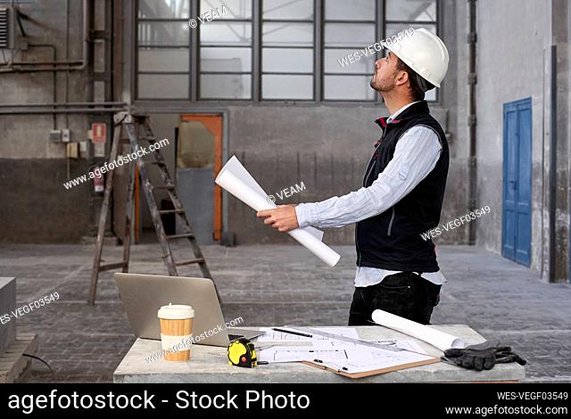 Male architect holding blueprint looking up while standing in building