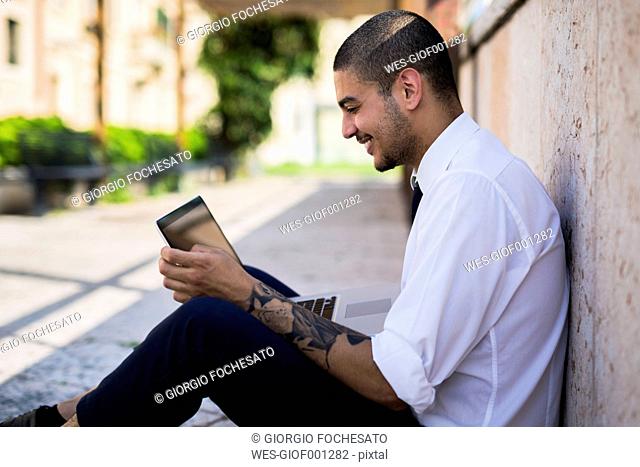 Smiling young businessman sitting on the ground looking at his tablet