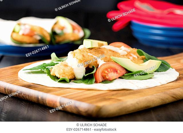 Crumbed fish fillet burrito with avocado and tomato serves on wooden cheese platter with rustic background
