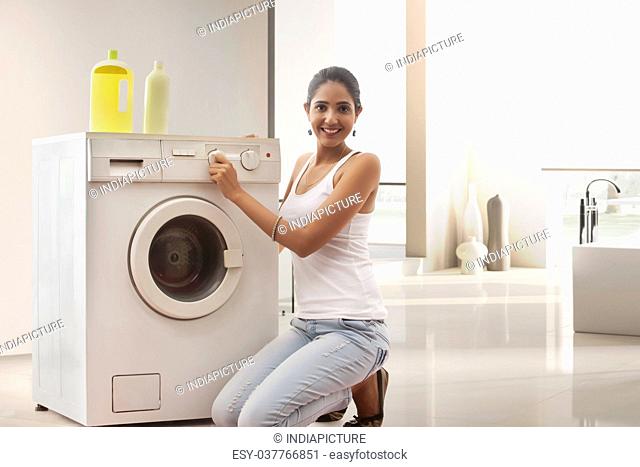 Smiling young woman turning washing machine dial in laundry room