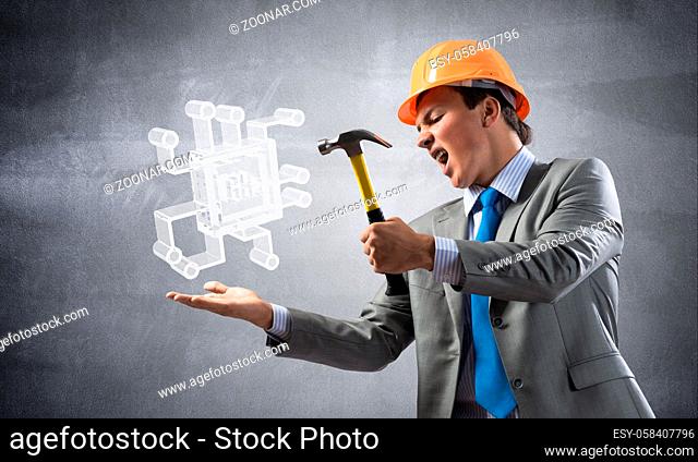 Furious businessman going to crash glass electronic microchip with hammer. Young man in business suit and safety helmet standing on wall background