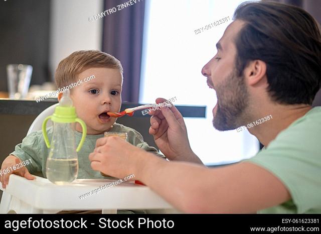 Breakfast for a babyboy. Young father feeding his cute baby son and looking funny