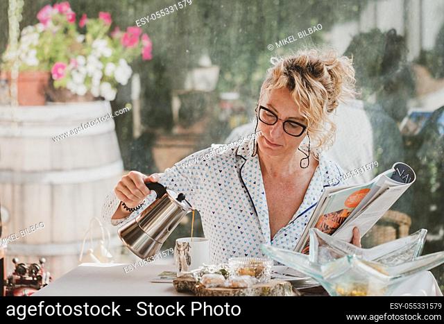 Blonde young mature woman with glasses in pyjamas at home in breakfast time, reading a magazine and preparing a cup of coffee