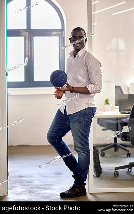 Smiling male professional with basketball leaning on doorway at office