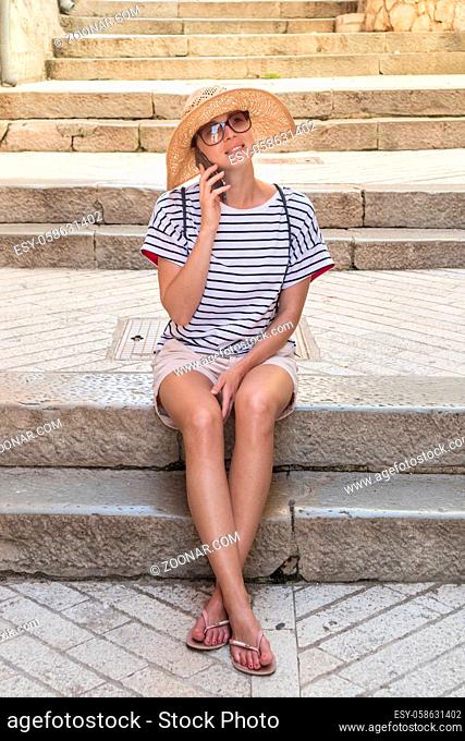 Beautiful young tourist woman at summer vacations, sitting on old stone steps of old medieval Mediterranean costal town, smiling