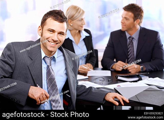 Smiling businessman looking at camera sitting at meeting table, coworkers in background